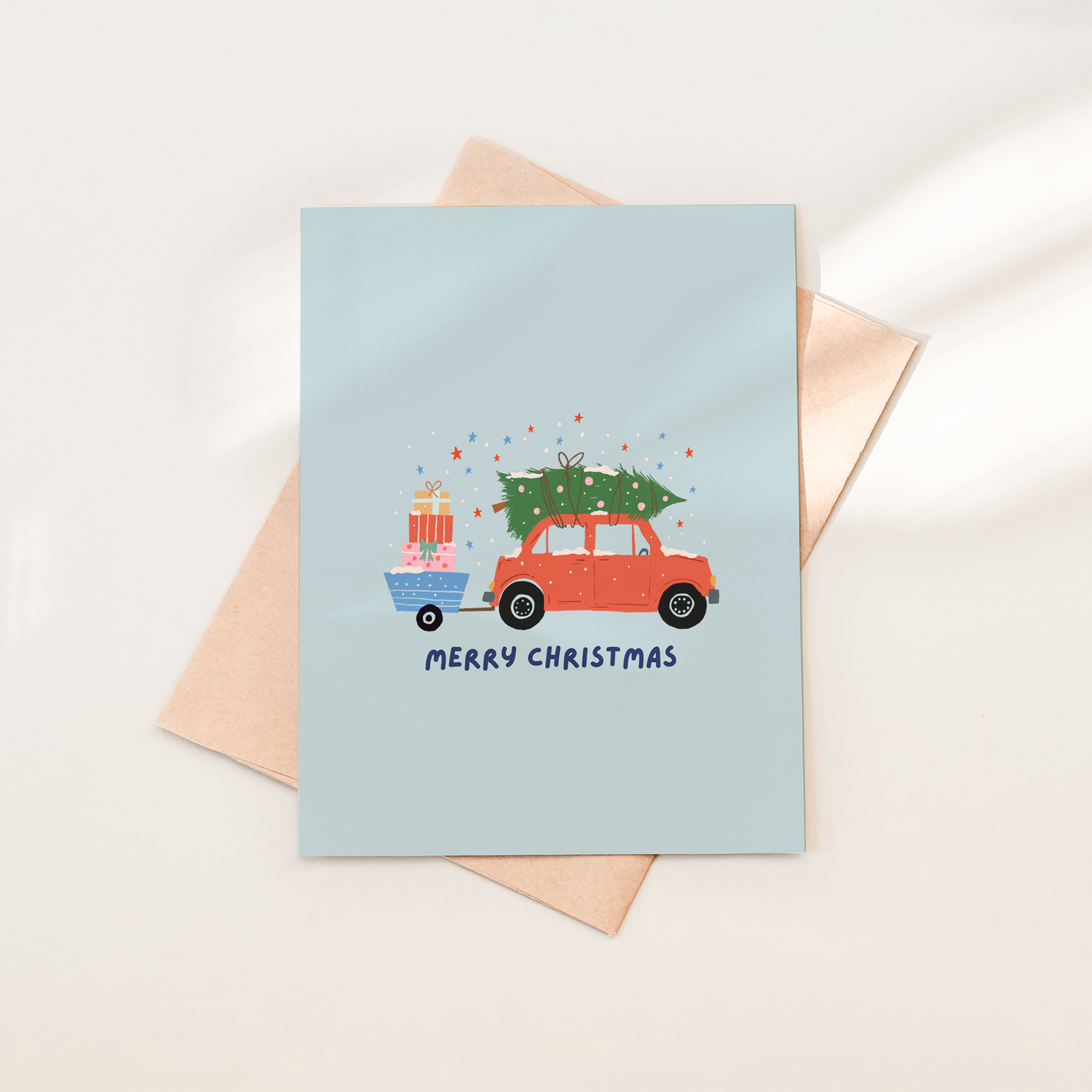 Christmas card with a red car and tree design on a light blue background, placed on a beige envelope.