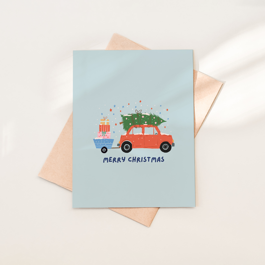 Christmas card with a red car and tree design on a light blue background, placed on a beige envelope.